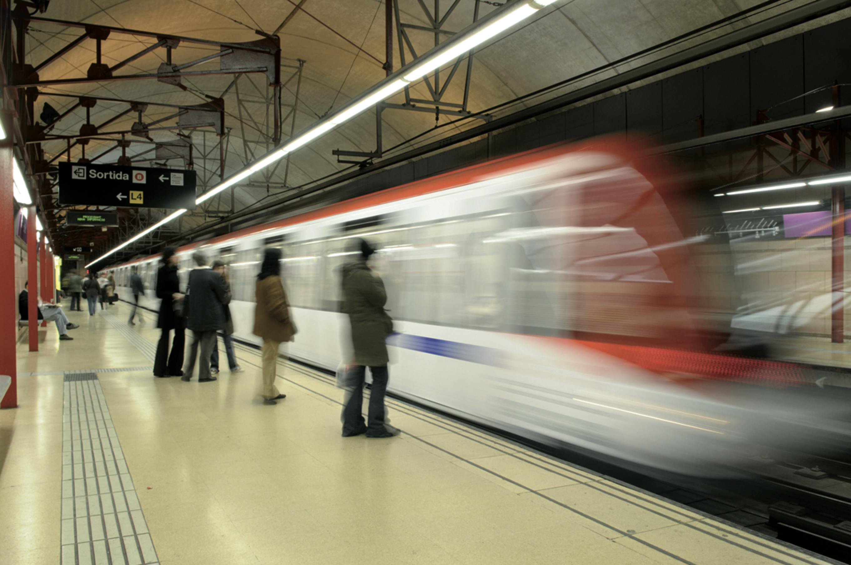 Pendolari in attesa su una banchina della metropolitana mentre un treno in rapido movimento passa sotto un alto soffitto industriale.