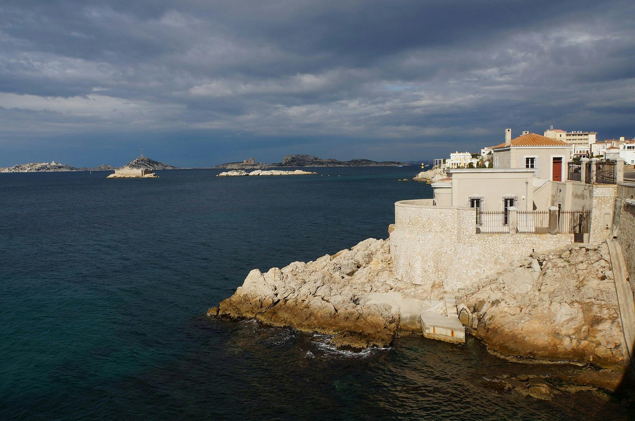 Rocky coastline with a stone building overlooking the sea; islands and a cloudy sky in the background.