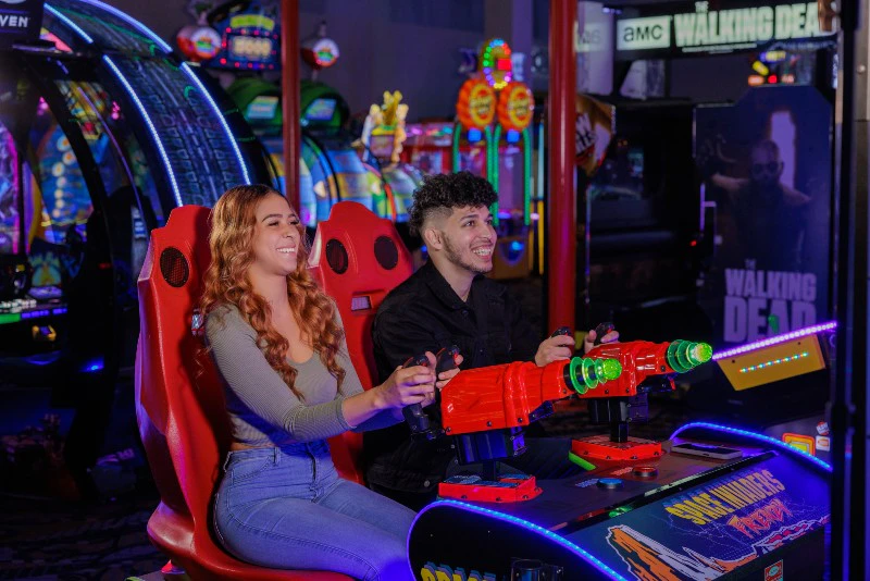 Two people smiling while playing an arcade game with red and green guns, surrounded by colorful arcade machines.