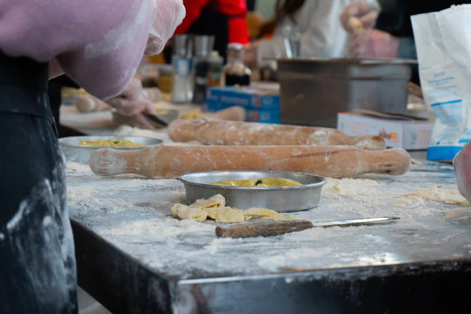 A busy kitchen scene with people preparing food, rolling pins, and baking tins filled with dough on a floured surface.