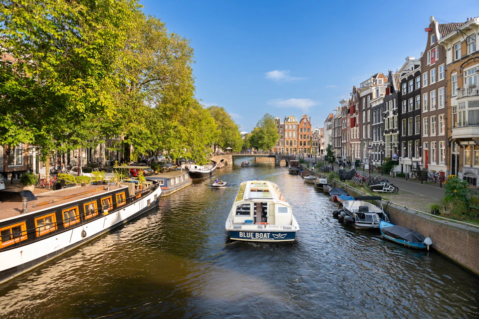A canal in a city with a tour boat labeled 'Blue Boat' surrounded by traditional buildings and trees under a blue sky.