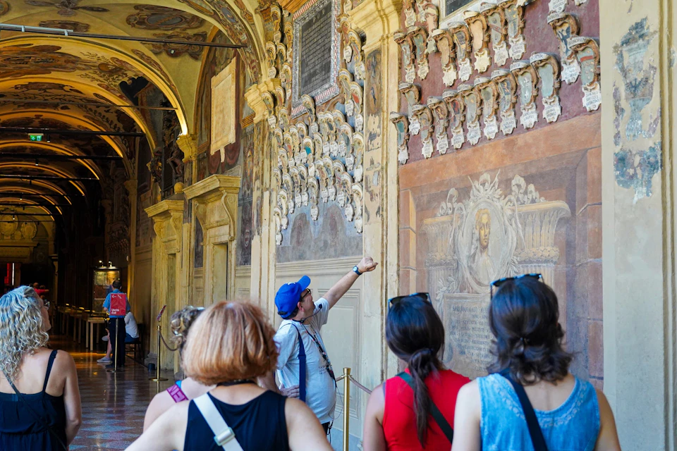A tour group looks at intricate wall carvings and murals inside an ornate historical building. The guide points at details.