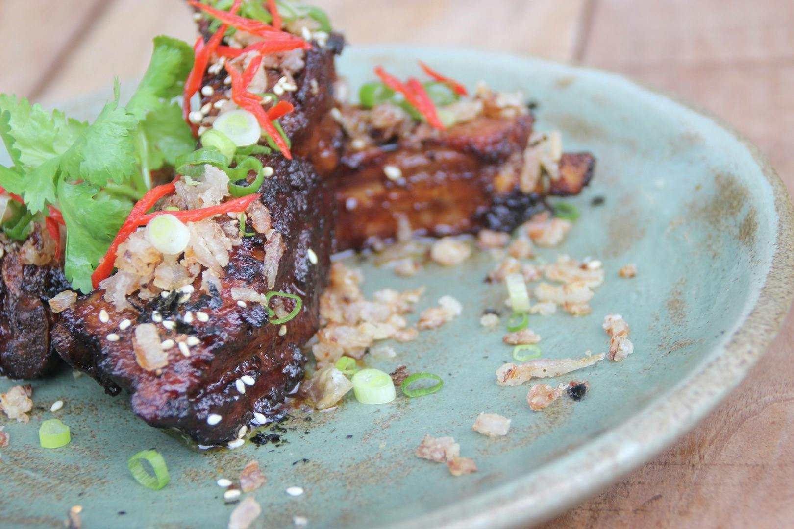 A plate of glazed ribs garnished with green onions, sesame seeds, red chili slices, and herbs on a green ceramic dish.
