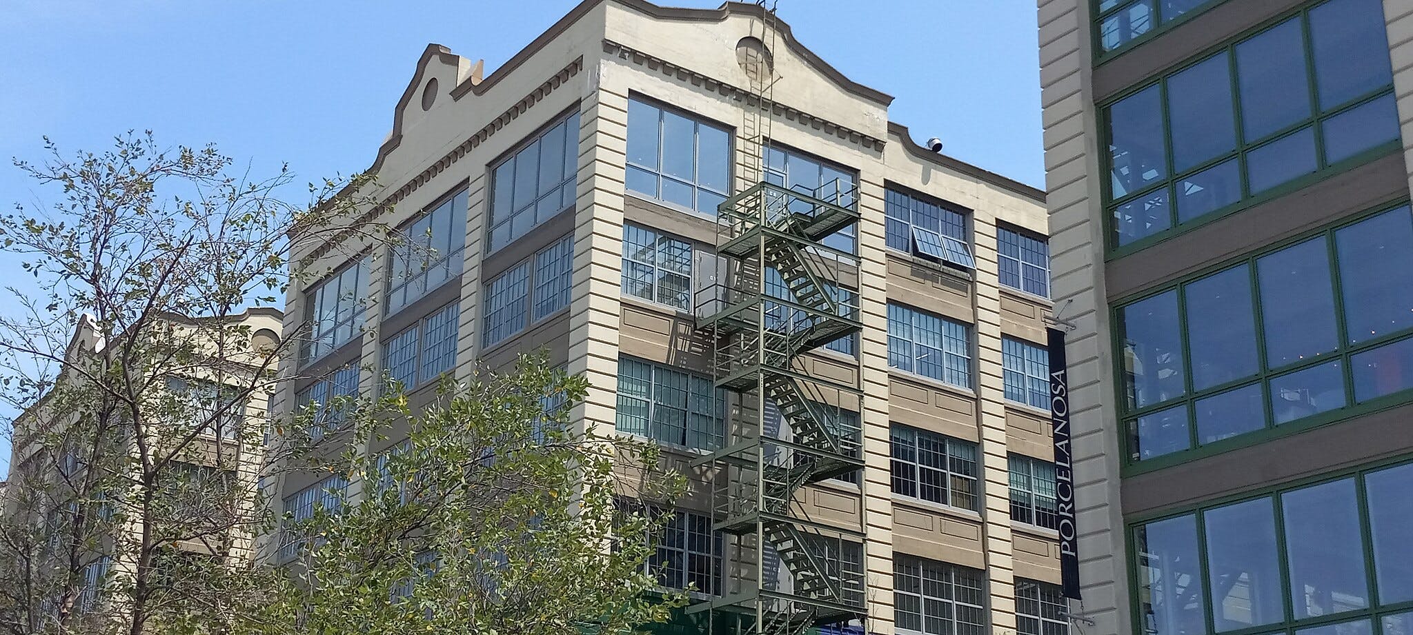 Edifício de vários andares com grandes janelas, escada de incêndio exterior e uma faixa com a inscrição "PORCELANOSA" visível. Ramos de árvores em primeiro plano.