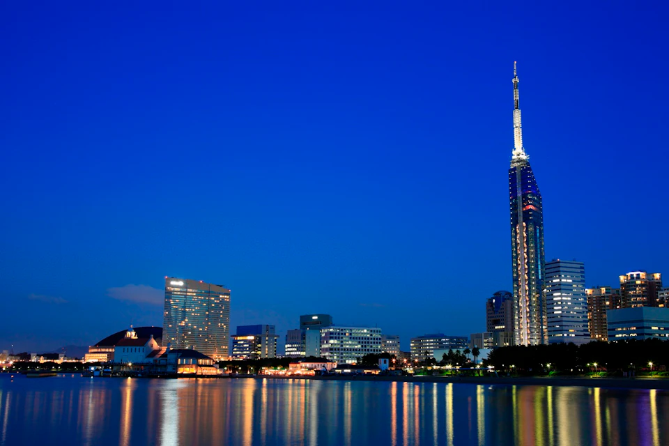 A cityscape at night featuring illuminated buildings and their reflections on a calm body of water under a clear sky.
