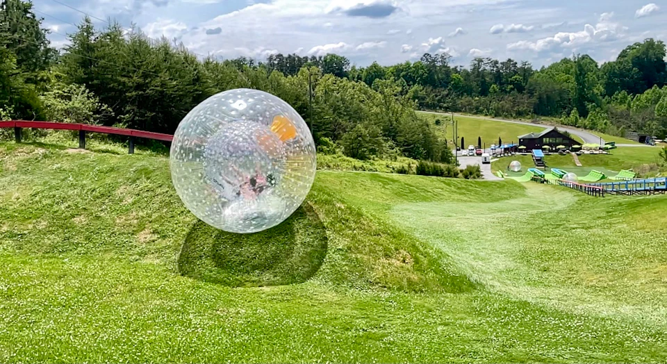 A person rolls down a grassy hill inside a large transparent zorb ball on a sunny day with trees and a road in the background.