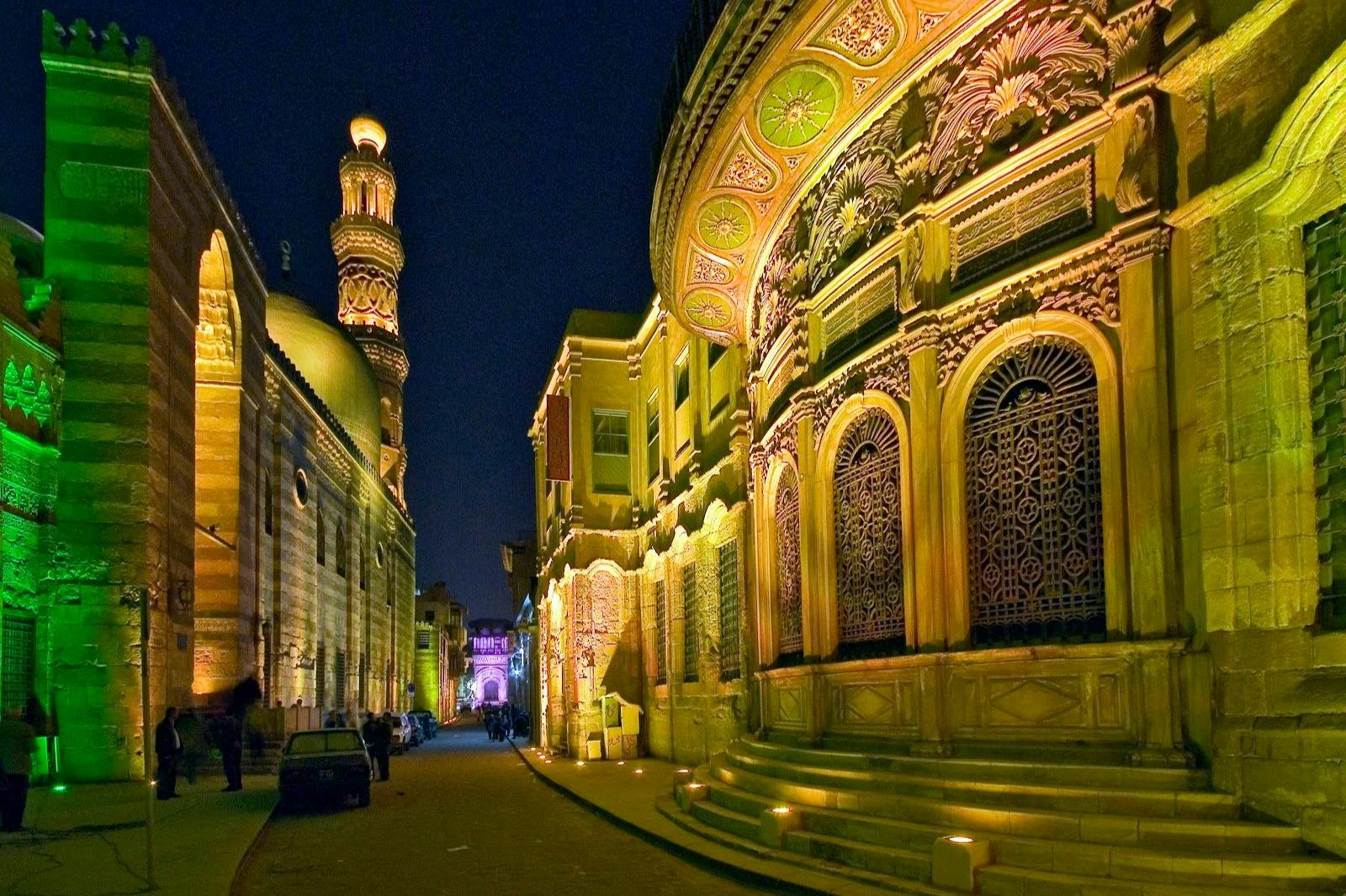 A well-lit, ornate historical street at night, with decorative façades and a minaret in the background.