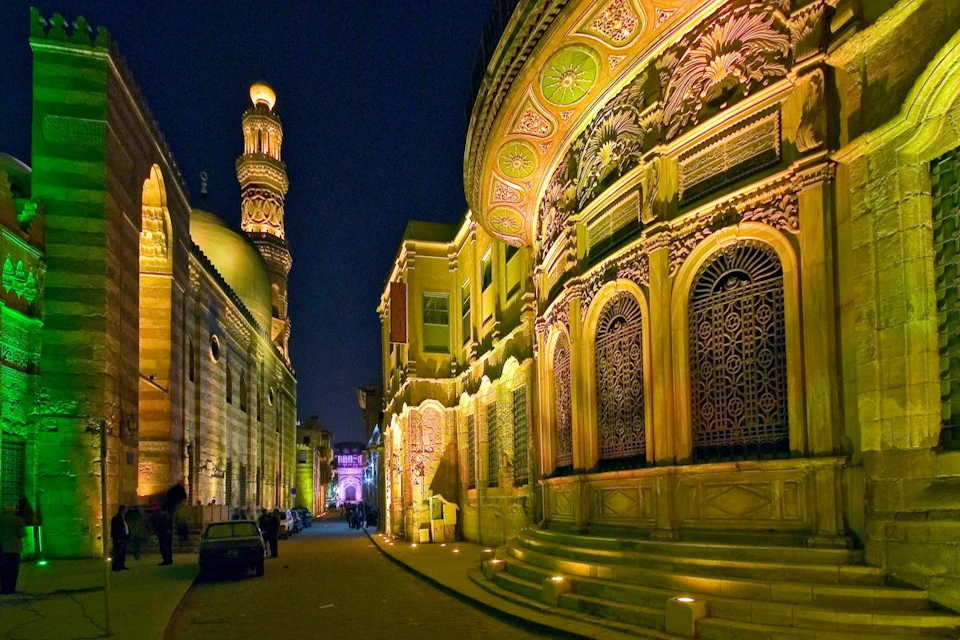 A well-lit, ornate historical street at night, with decorative façades and a minaret in the background.