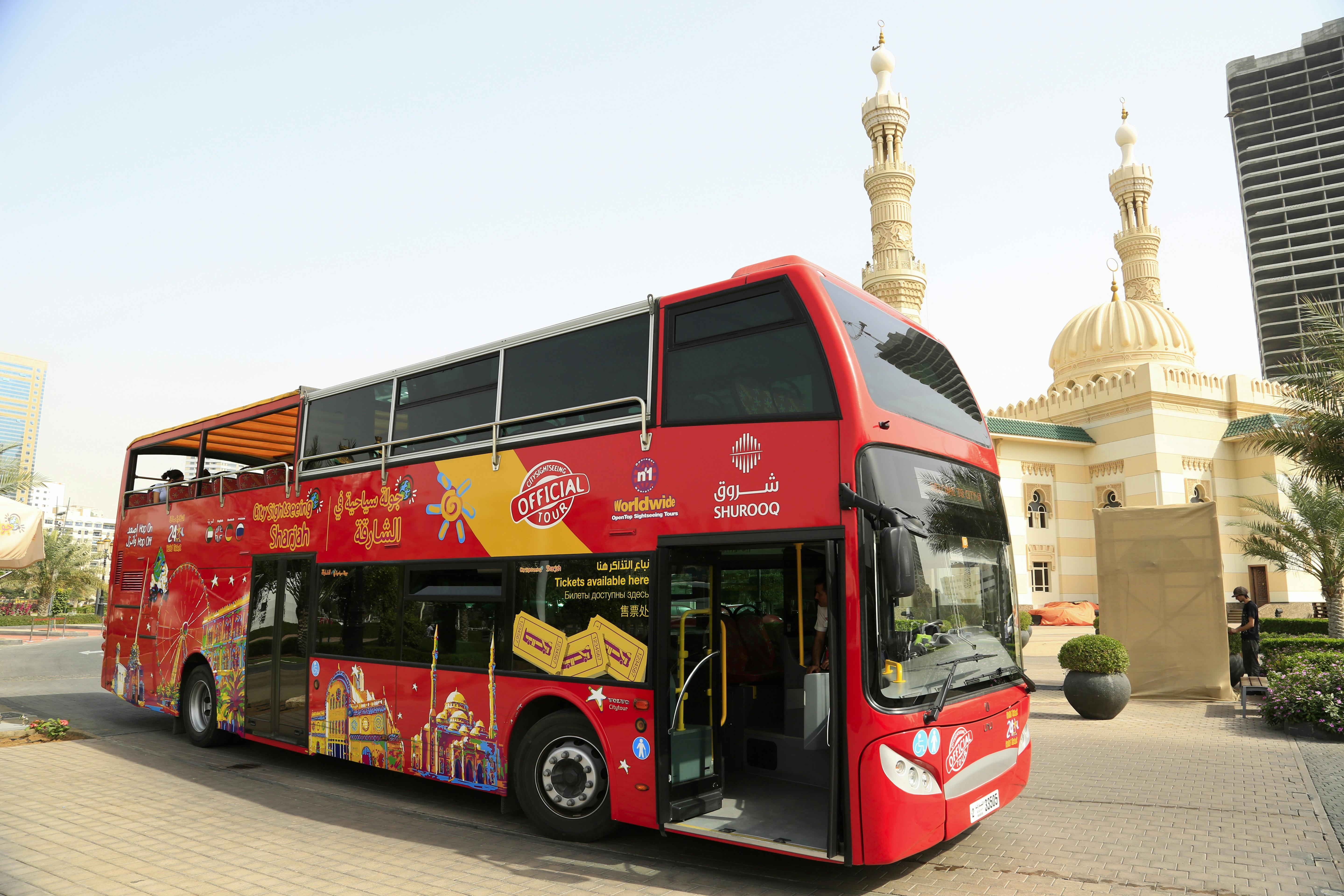 Red double-decker tour bus parked near a building with domes and a tall minaret under a clear sky.