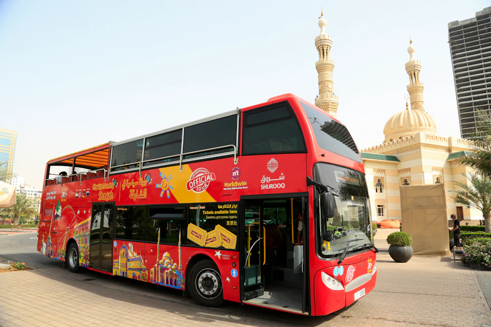Red double-decker tour bus parked near a building with domes and a tall minaret under a clear sky.