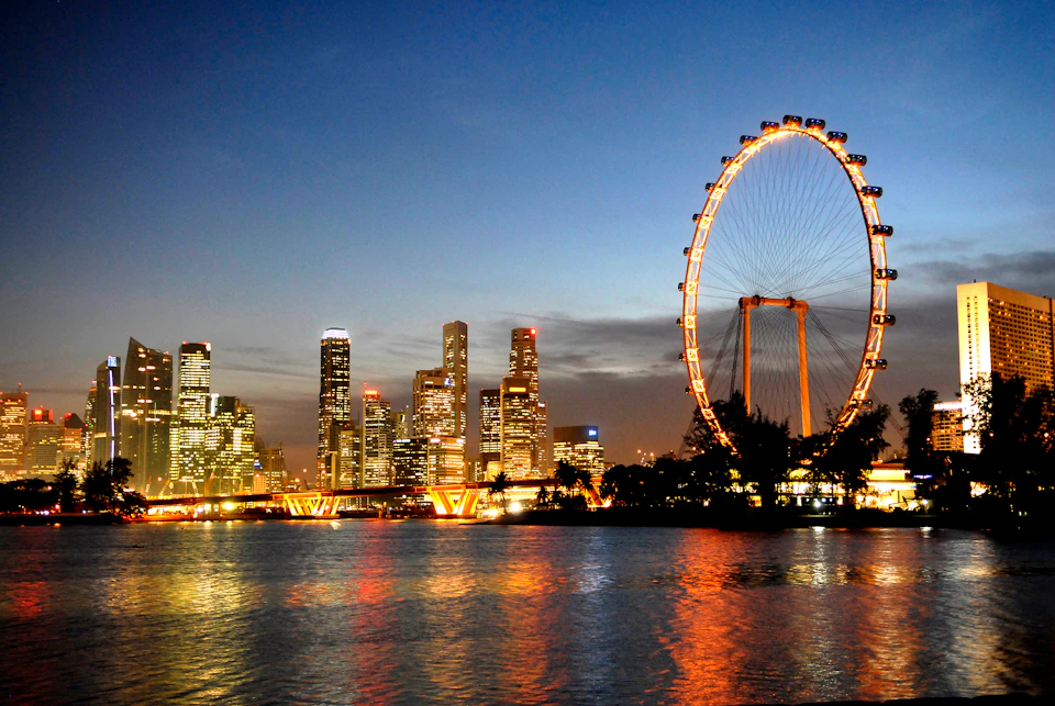 City skyline with illuminated skyscrapers at dusk, a large lit Ferris wheel, and reflections on a calm body of water.