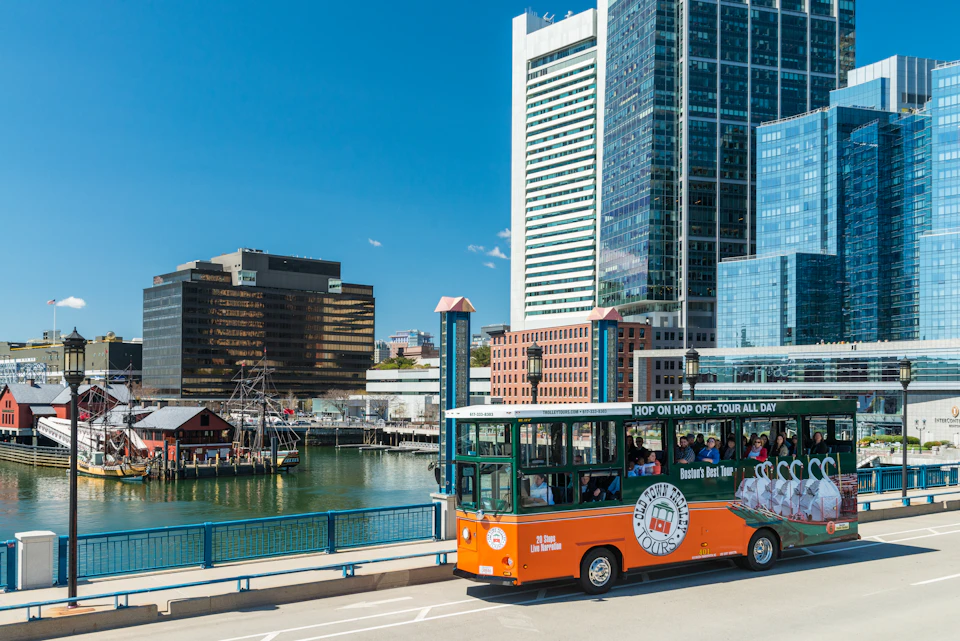 An orange tour bus drives past waterfront buildings and a ship, with tall modern skyscrapers in the background.