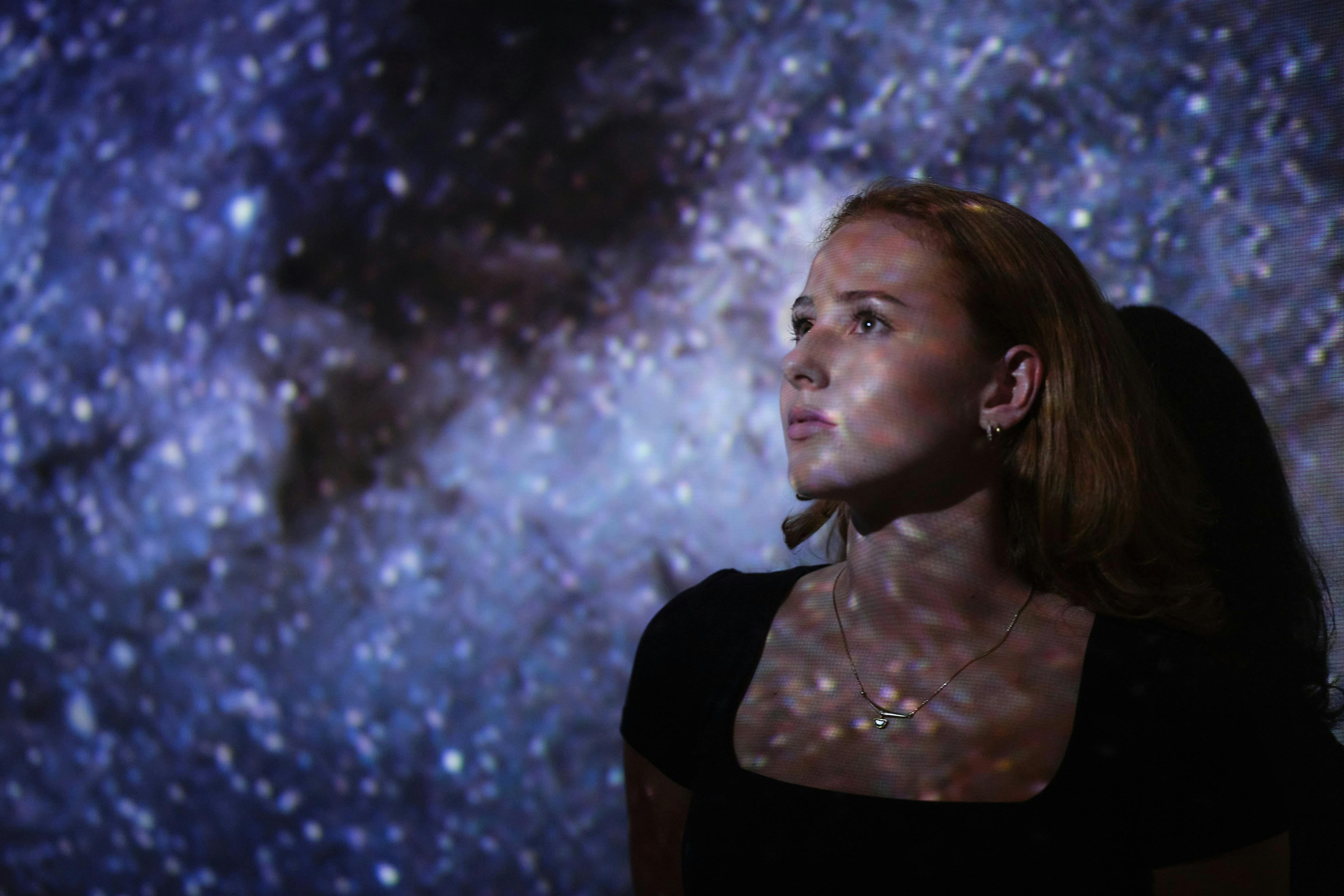 En kvinna i en mörk outfit belyses av en stjärnklar projektion, som tittar upp mot en kosmisk bakgrund.