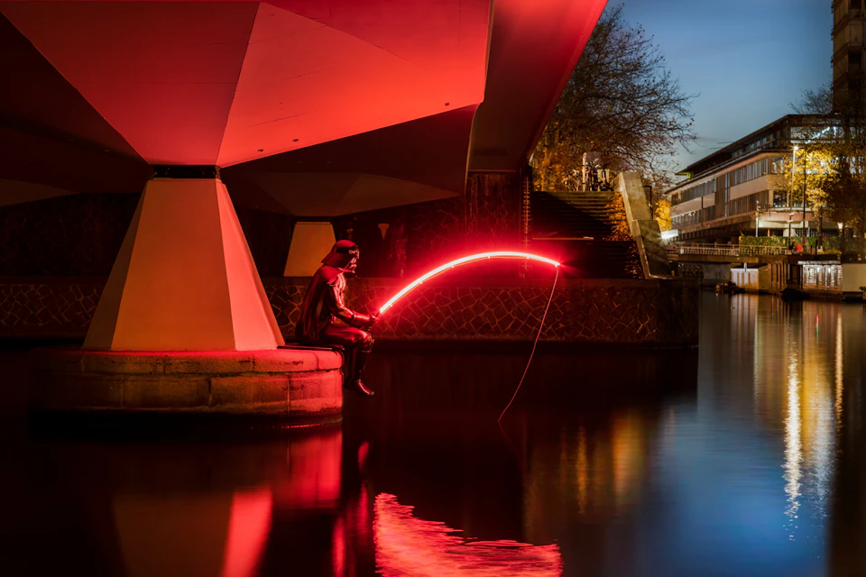 A person in a costume sits by a canal at night, holding a glowing red fishing rod under a bridge.