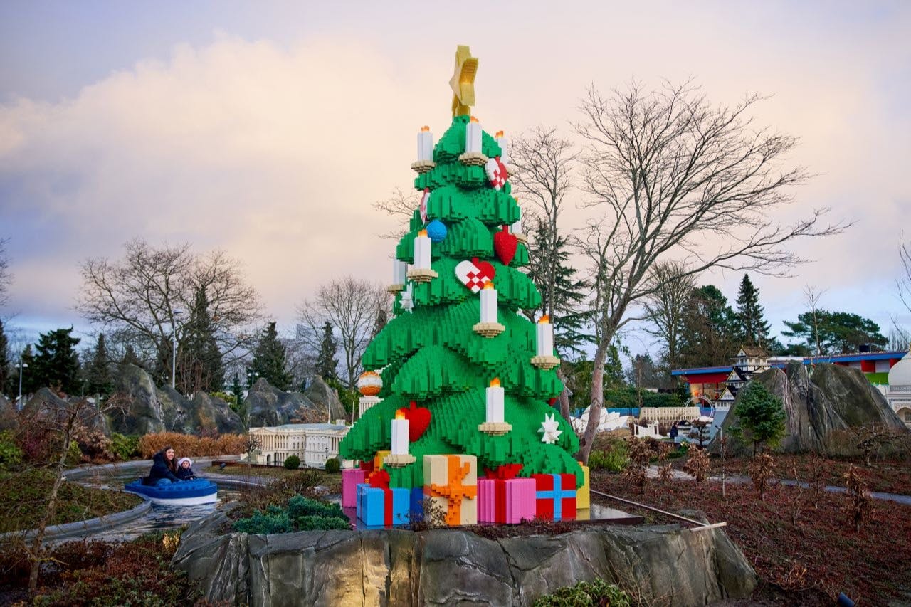 Большая рождественская елка LEGO, украшенная свечами и орнаментами, в окружении обернутых подарков. Два человека катаются на лодке неподалеку.