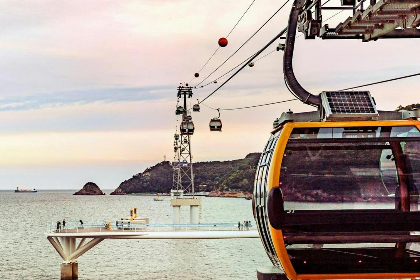 Cable cars traveling above water with a distant shoreline and hills under a pastel-colored sky.