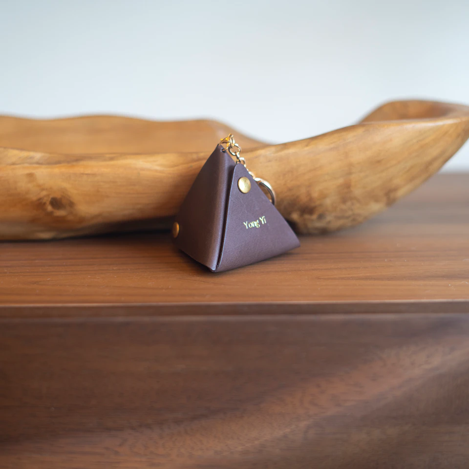 A brown triangular key pouch with gold details and a keychain, placed on a wooden surface near a carved wooden bowl.