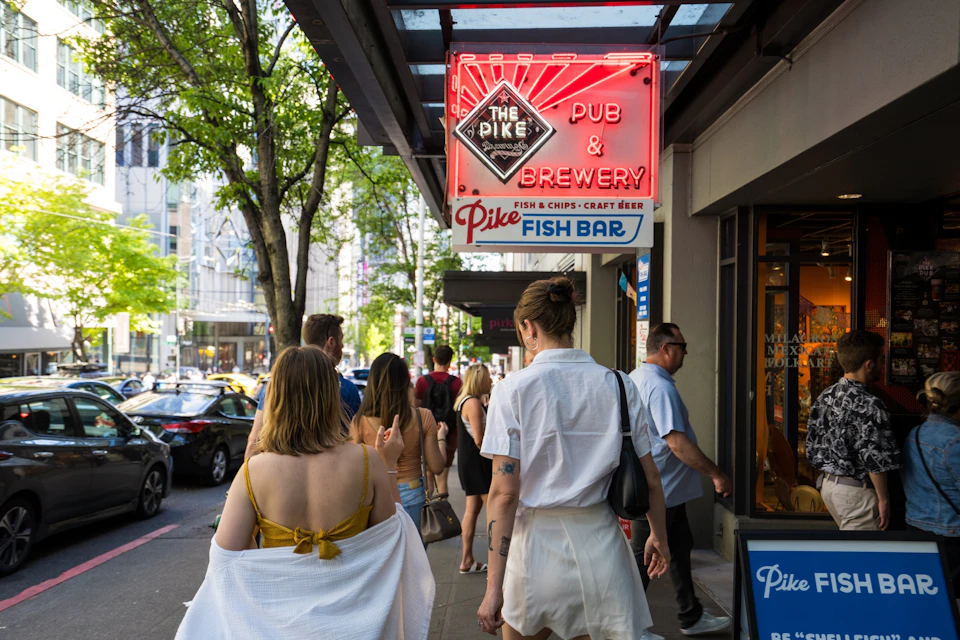 People walking on a city sidewalk past a pub and brewery with a neon sign reading "The Pike Pub & Brewery."