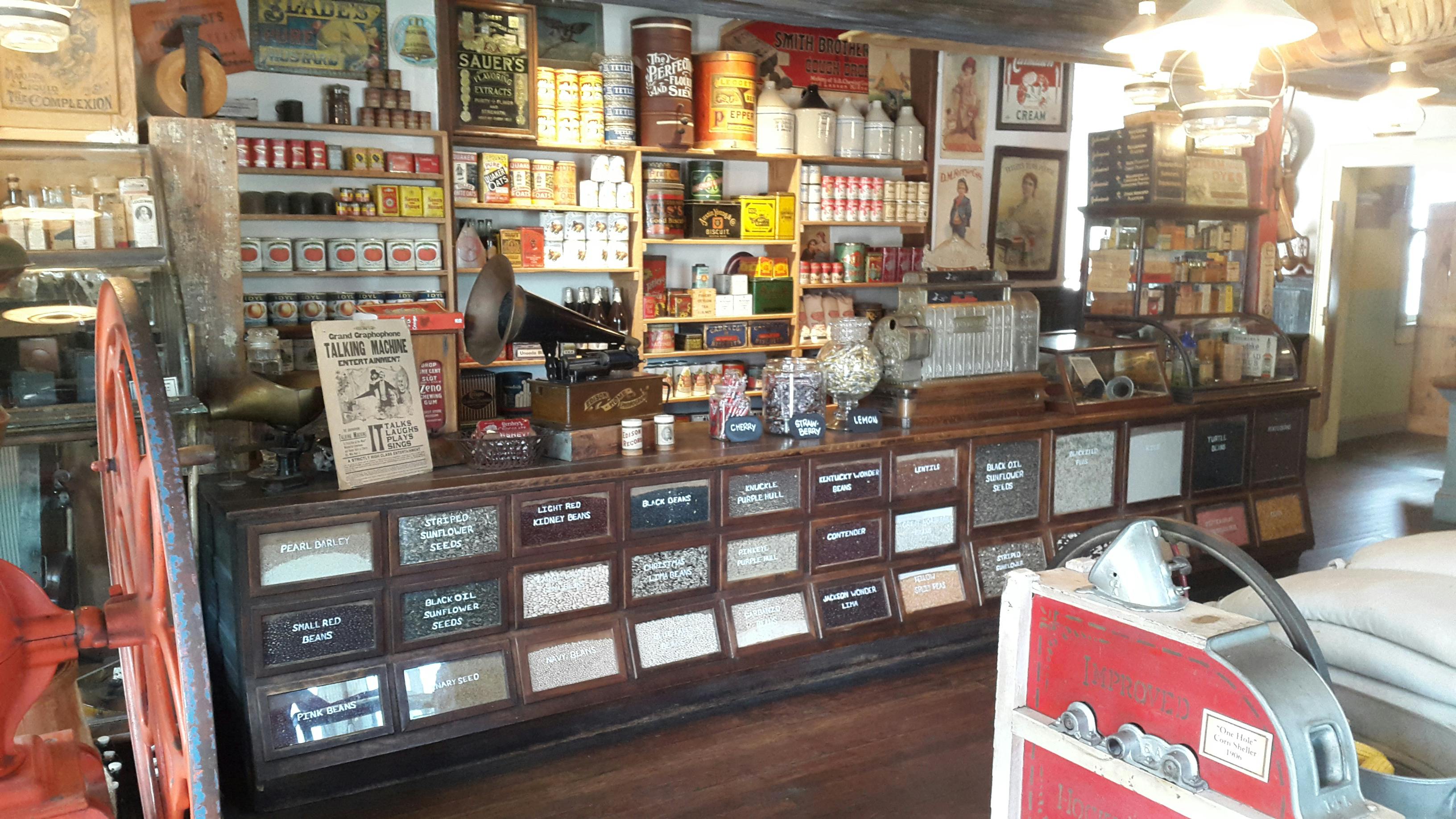 Ein altmodischer Gemischtwarenladen mit Holzregalen, die mit Konserven, Gewürzen und Gläsern gefüllt sind. Auf dem Tresen steht ein altes Grammophon.