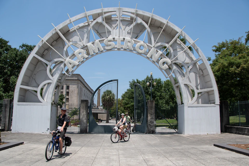 Cyclists ride past an arched entrance labeled "ARMSTRONG" in a park setting on a sunny day.