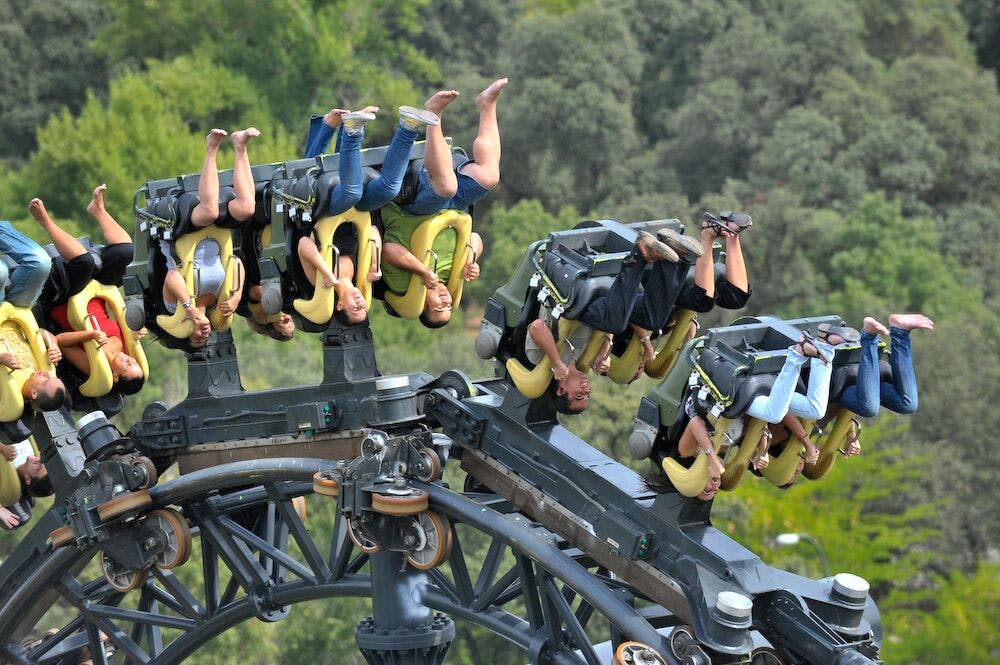 人们倒立在环形过山车上，坐在森林背景下，坐在黄色座椅和复杂的金属支架上。