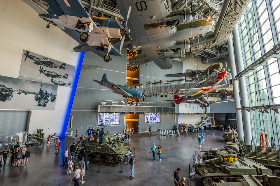 People exploring an aircraft and military exhibit indoors, with planes suspended above and military vehicles on the ground.