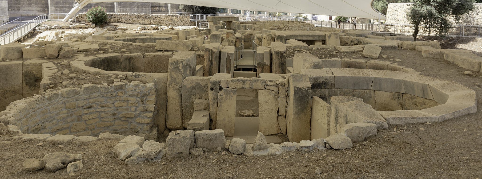 Antigas ruínas de pedra com câmaras circulares e portas rectangulares, rodeadas de terra e paredes parciais de pedra sob um dossel.