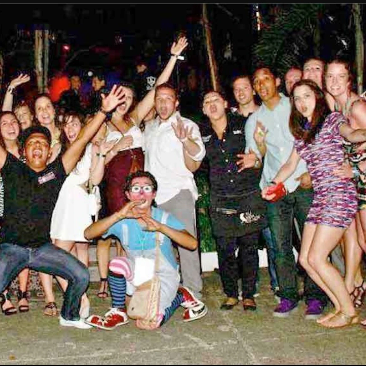 A group of people posing together outdoors at night, some with expressive gestures and colorful attire, smiling towards the camera.