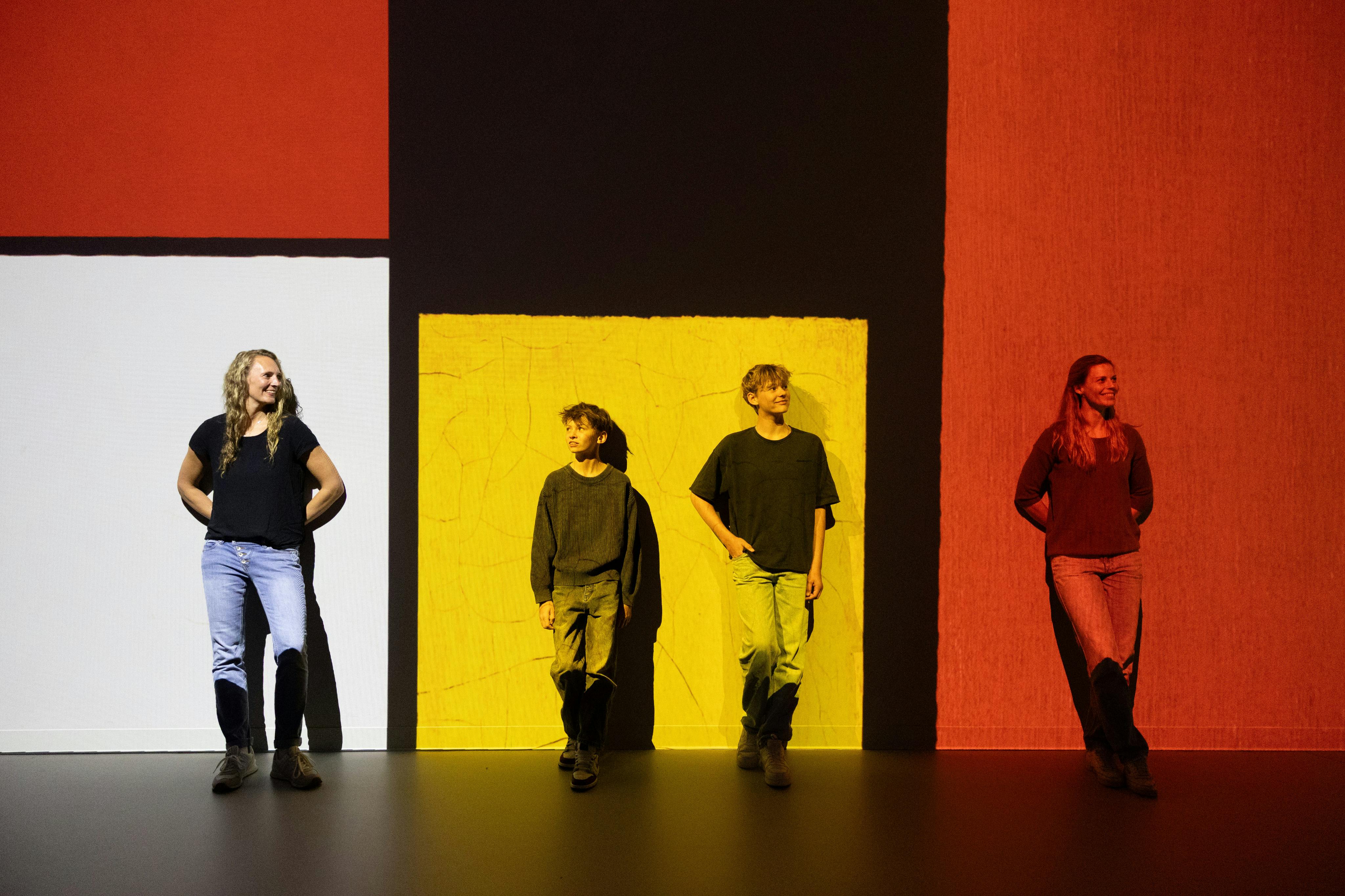 Three people stand in front of a colorful geometric art installation with red, white, and yellow panels on a black wall.