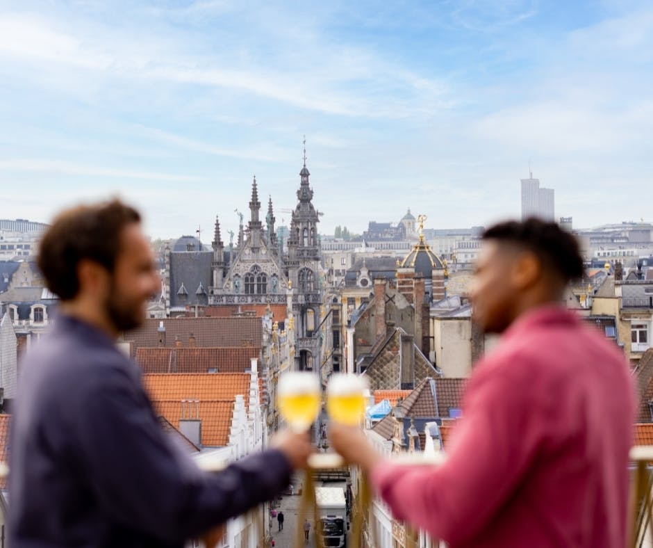 Persone sedute su una terrazza sul tetto al tramonto, sotto le luci dei fili, che bevono un drink con il paesaggio urbano sullo sfondo.