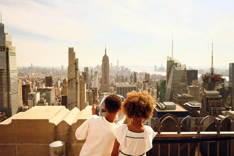 Two children stand on a rooftop observation deck, looking at skyscrapers through a coin-operated viewer on a sunny day.