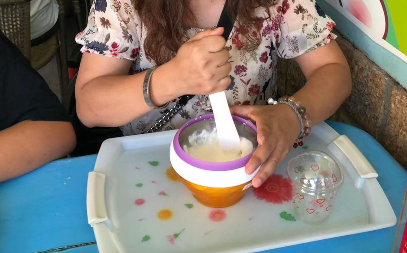 A person stirring ingredients in a bowl on a tray with a floral design, next to a transparent cup with heart patterns.