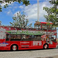 Röd, öppen turnébuss som främjar Liverpool Football Club-stadion turer, parkerad på en kullerstensgata med träd i bakgrunden.