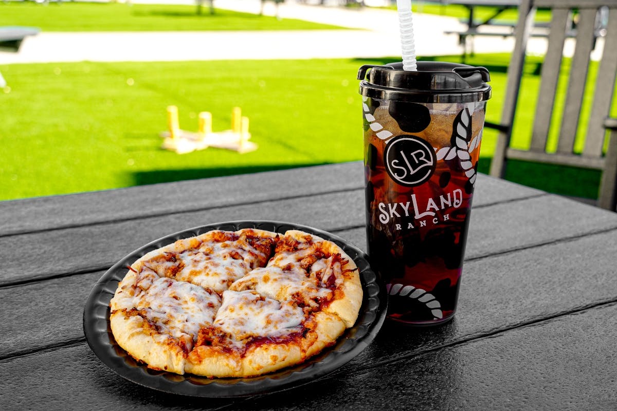 Personal pizza on a black plate next to a large drink cup with a straw, labeled "SkyLand Ranch," on a picnic table with grass in the background.