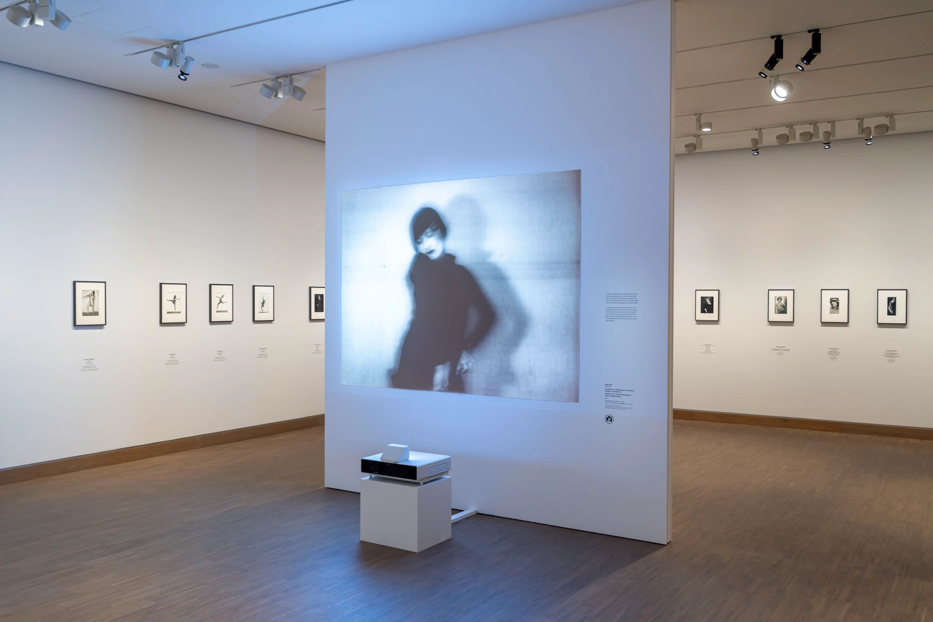 An art gallery with a projection of a blurred figure on a wall, surrounded by framed photographs and a projector on a stand.