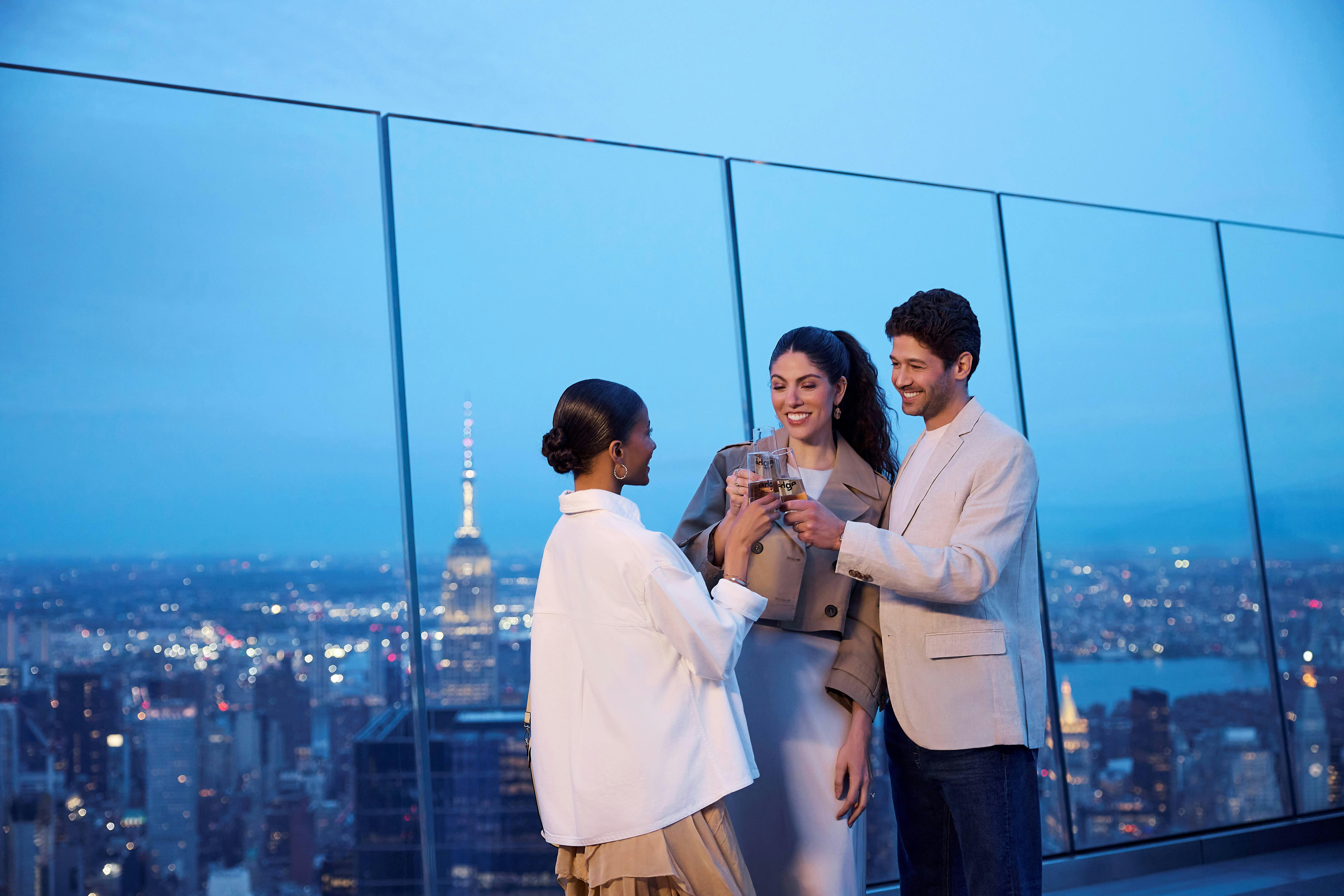 Tři lidé si připíjejí na střeše s panoramatem města a osvětlenými budovami v pozadí.