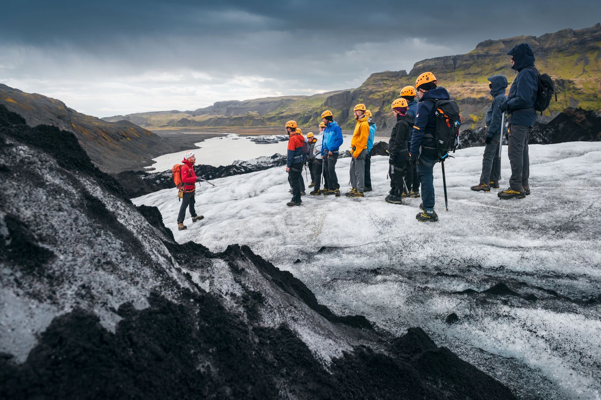 A group of people wearing helmets and outdoor gear stand on a glacier, with rocky terrain and hills in the background.