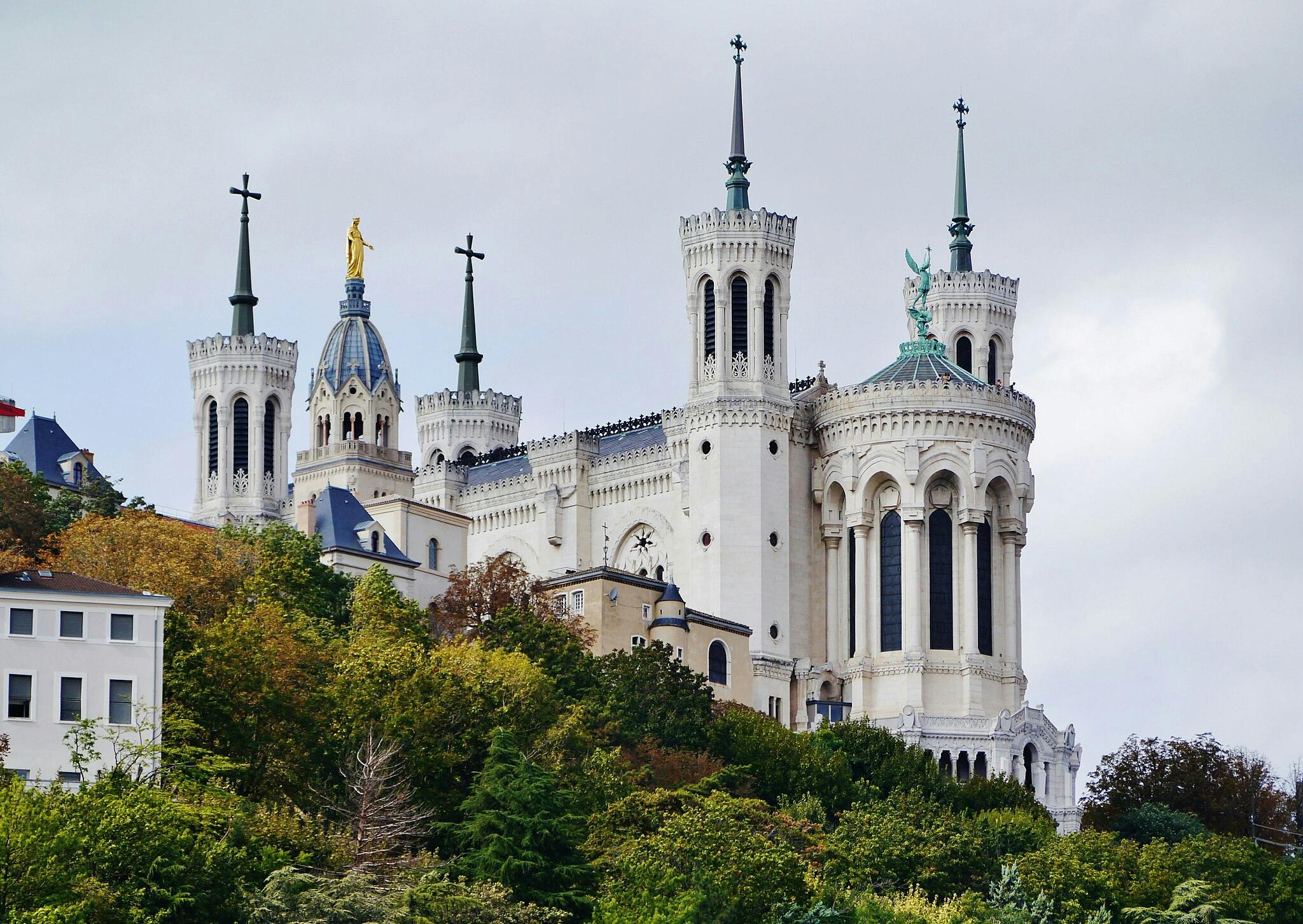Na zalesněném svahu pod zataženou oblohou stojí velká, zdobená katedrála se čtyřmi věžemi, zakončená kříži a sochami.