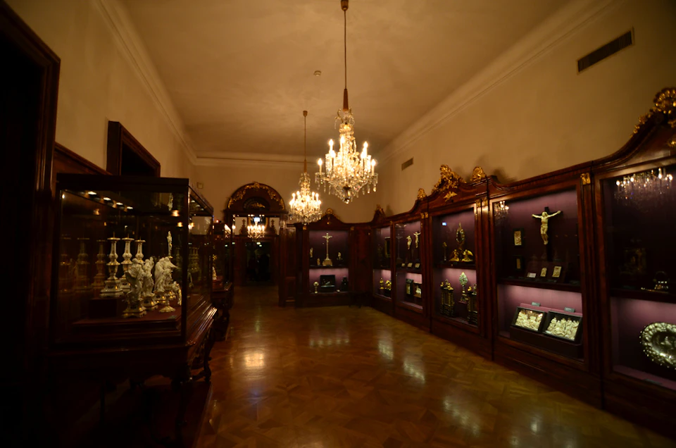 Ornate room with chandeliers, showcasing religious artifacts in wooden display cases with purple lighting. Polished wooden floor.