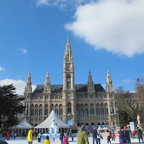 Folk åker skridskor framför en stor byggnad i gotisk stil med höga spiror, under en klarblå himmel.