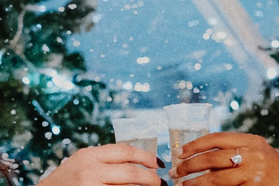 Two people in winter attire toast with drinks while seated inside a festive, snow-covered dome decorated with Christmas trees.