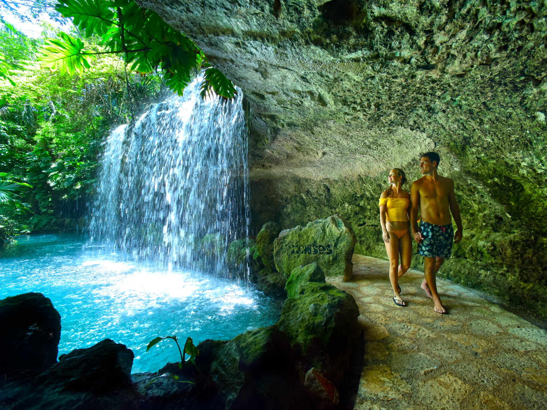 A couple walks on a stone path near a waterfall inside a cave, with lush greenery visible overhead.