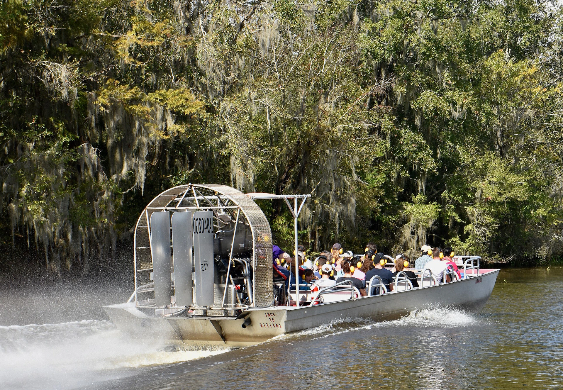 A group of people rides on an airboat through a tree-lined swamp area.