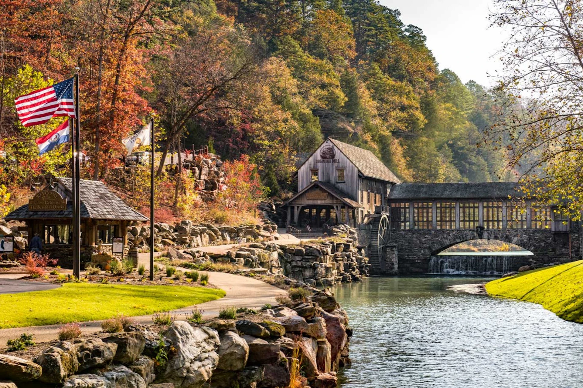 A rustic mill building with a water wheel next to a river, surrounded by autumn trees and rocky landscaping.