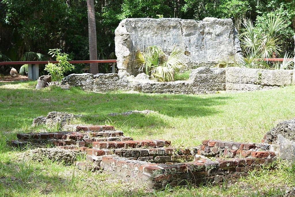 Ruins of a historic building surrounded by grass, trees, and plants, with remnants of brick and stone walls visible.