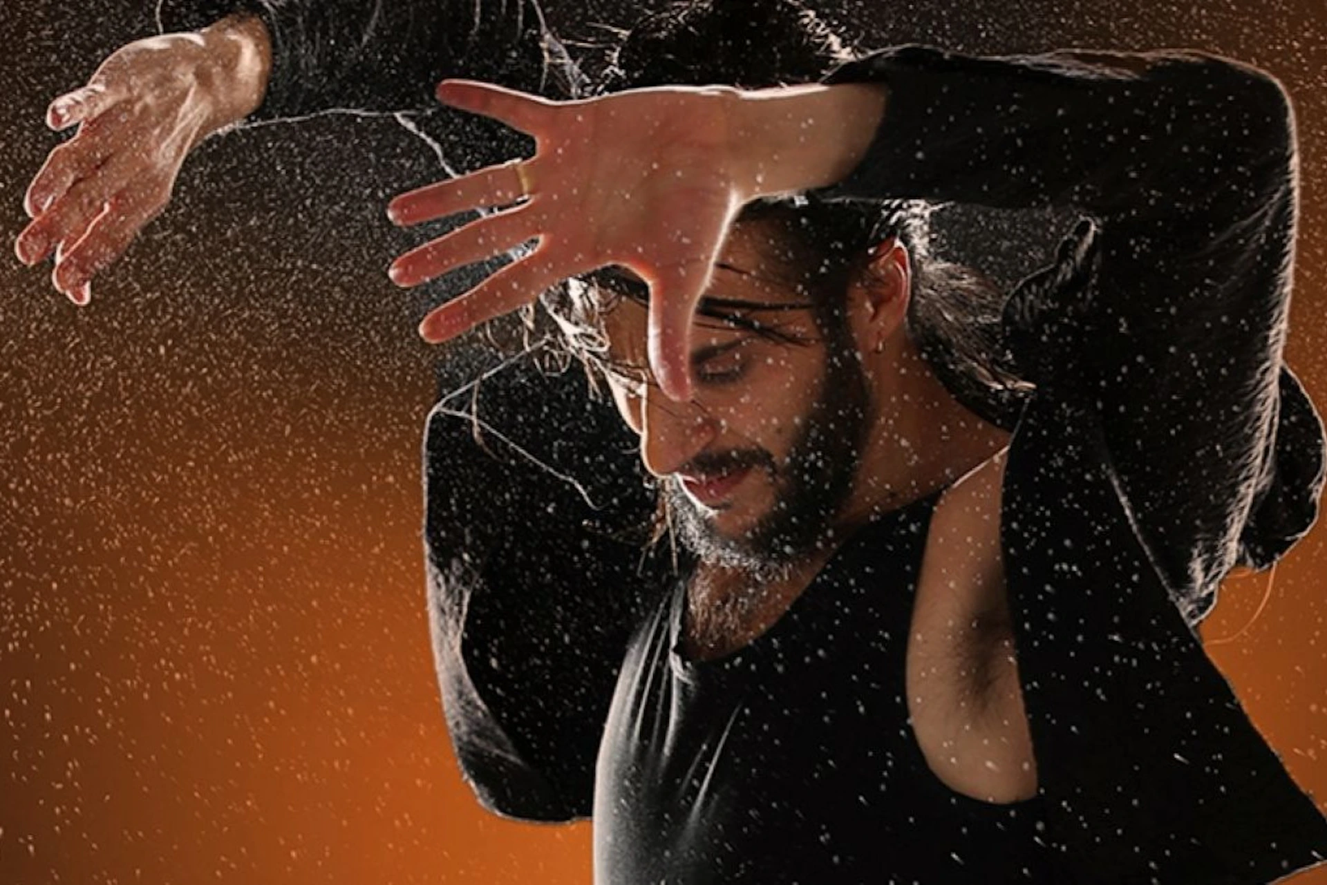 A person in a dynamic pose with water droplets around them, wearing a black outfit against an orange background.