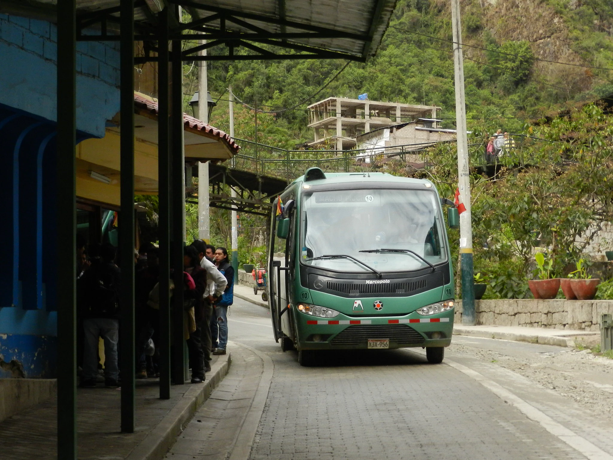 A green bus travels through a narrow, tree-lined street with people waiting at a covered stop on the left side.