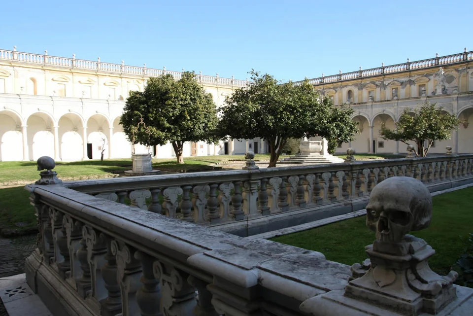 A courtyard with trees, statues, a balustrade, and historical architecture under a clear blue sky.