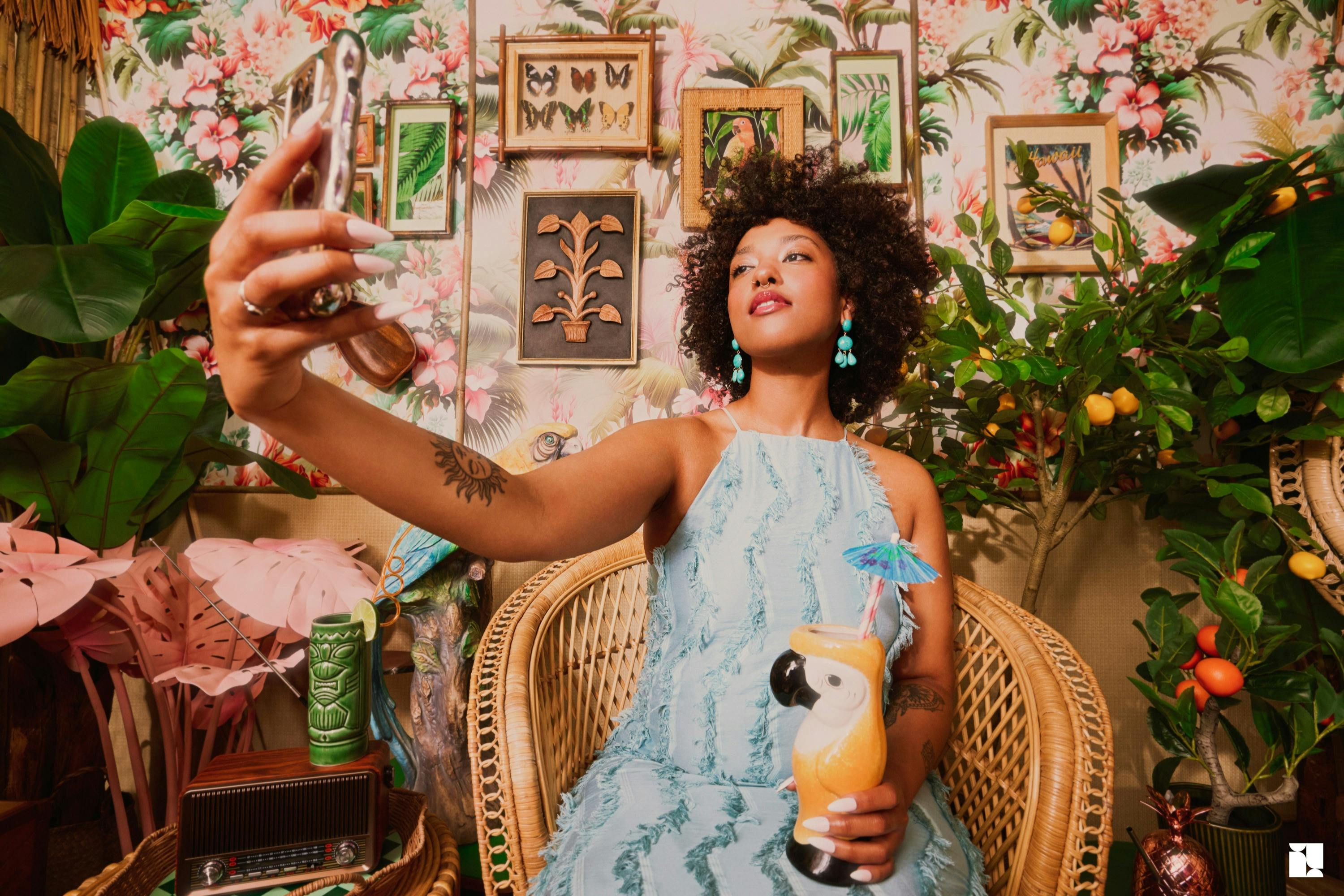 Ένα άτομο με μπλε φόρεμα κάθεται σε μια ψάθινη καρέκλα, κρατώντας ένα τροπικό ποτό, περιτριγυρισμένο από πολύχρωμη διακόσμηση και πράσινα φυτά.