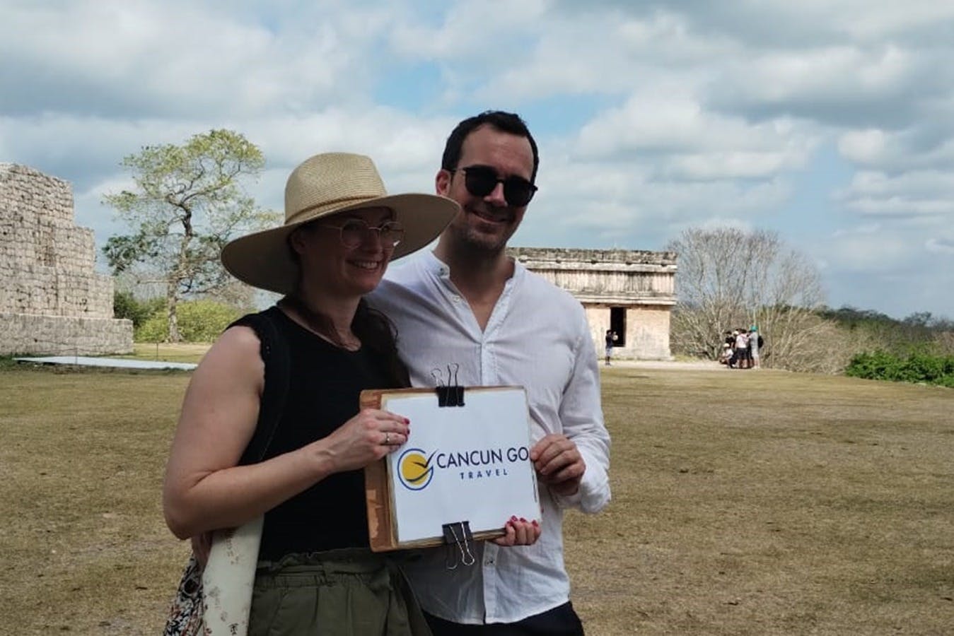 Deux personnes sourient dans un espace ouvert, tenant une pancarte sur laquelle on peut lire "Cancun Go Travel". Des ruines et des arbres sont visibles à l'arrière-plan.